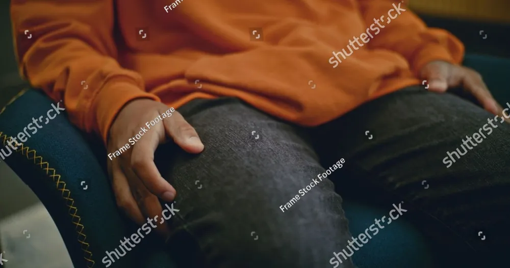 A person in an orange sweater sitting with a hand resting on one knee