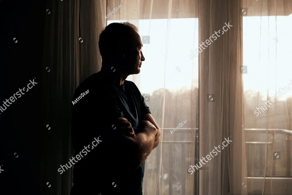 A man in silhouette at a bright window with his arms crossed