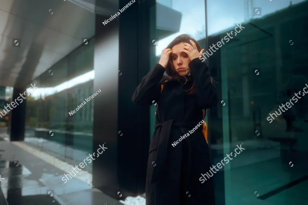 A woman in a dark coat holding her head in a busy glass hallway