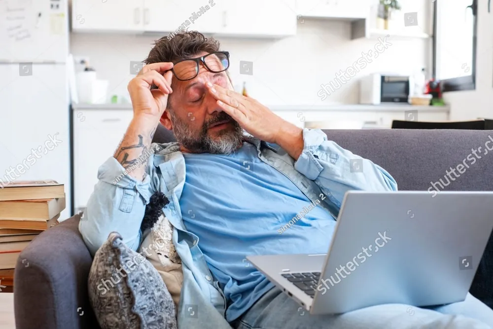 A tired man rubbing his eyes beside his laptop on the couch
