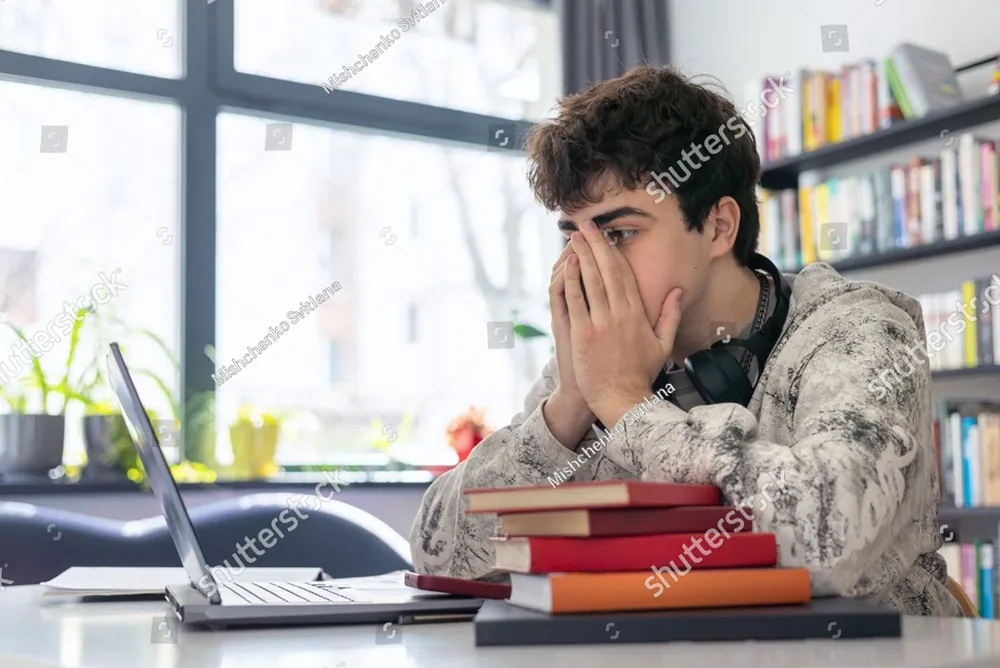 A teenager covering his face at a desk stacked with books